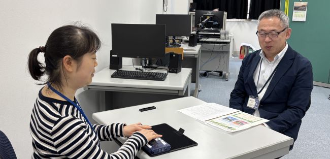 【写真】大阪府ITステーションの視覚障がい者講習室で行われたインタビューの様子。テーブルをはさんで対面に座っています。右手奥が中津サテライトオフィスの安藤さん、左手手前が幸田。