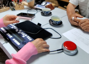 写真：
ボタンスイッチ型の音声録音再生装置（VOCA）にスイッチを付けられるようにしたもの。もともとボタンを押すと録音が再生される装置だが、左手のマイクロスイッチを押すことで録音が再生される。