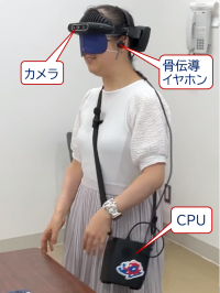 写真:SYNCREO（実験機）を装着した幸田。カメラと骨伝導イヤホンで構成された装着部と肩掛けするCPU部に分かれ、ケーブルでつています。